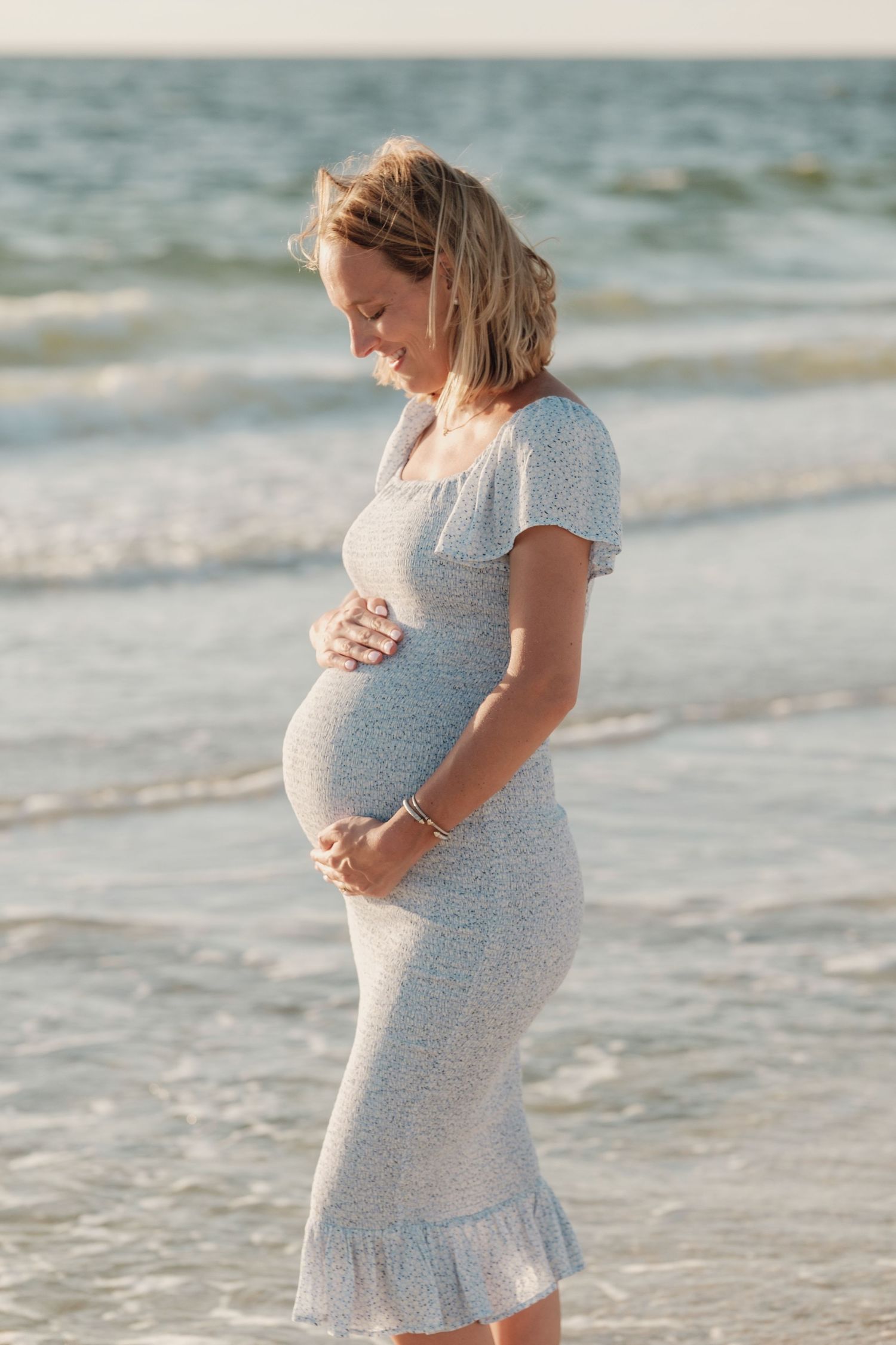 Expectant mother in a grey dress poses on the beach during golden hour.