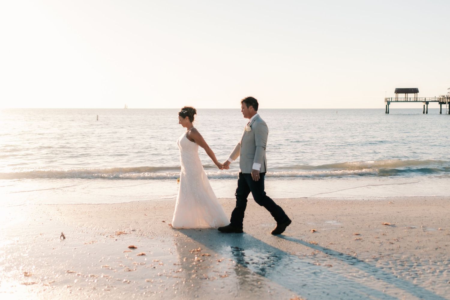 Bride and groom walk hand in hand along the shoreline at golden hour on their wedding day.