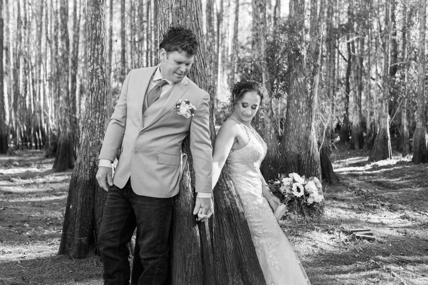 Black and white photo of bride and groom walking hand in hand through a forest of tall pine trees.