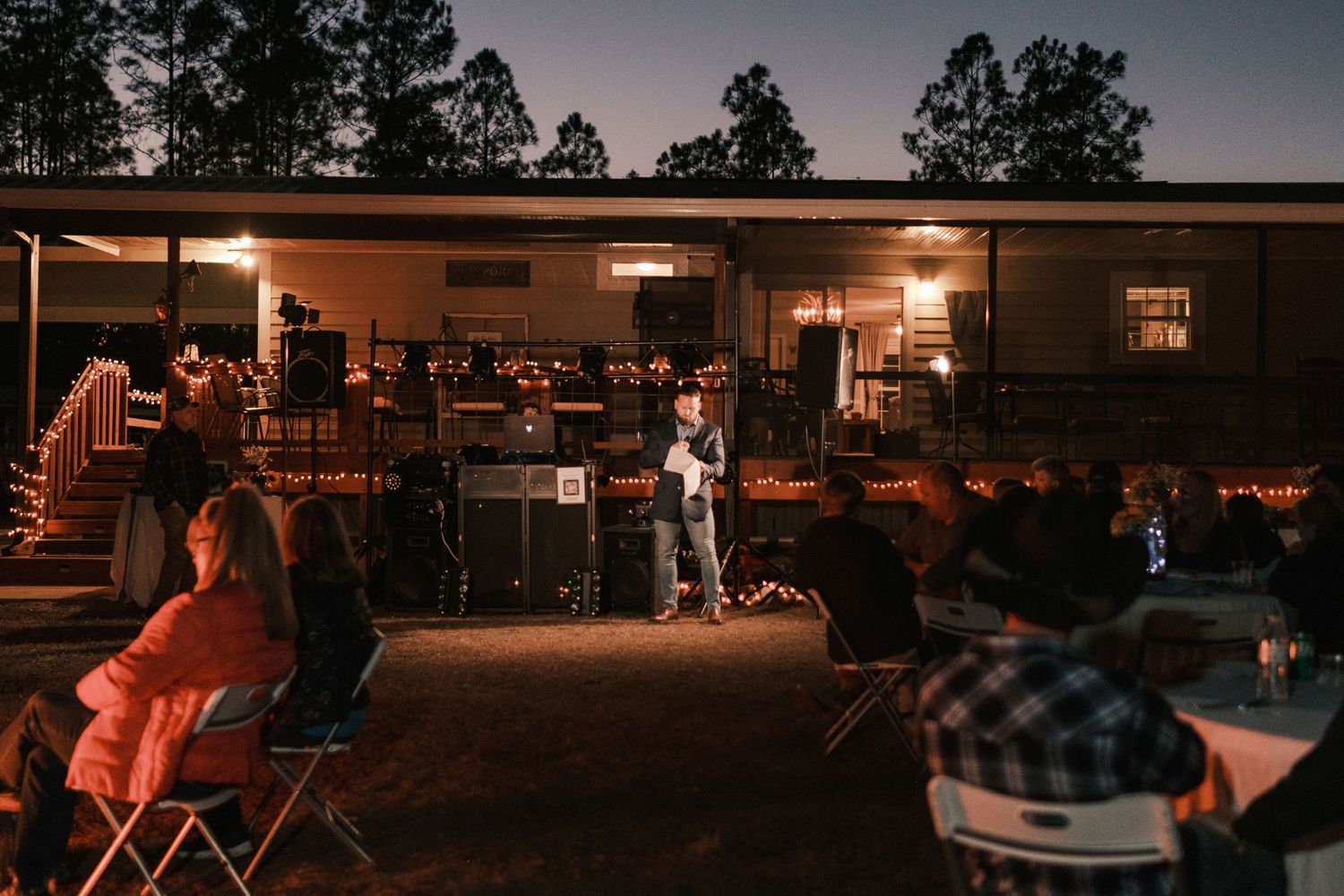 Outdoor evening wedding reception with guests seated as officiant speaks under warm string lights.