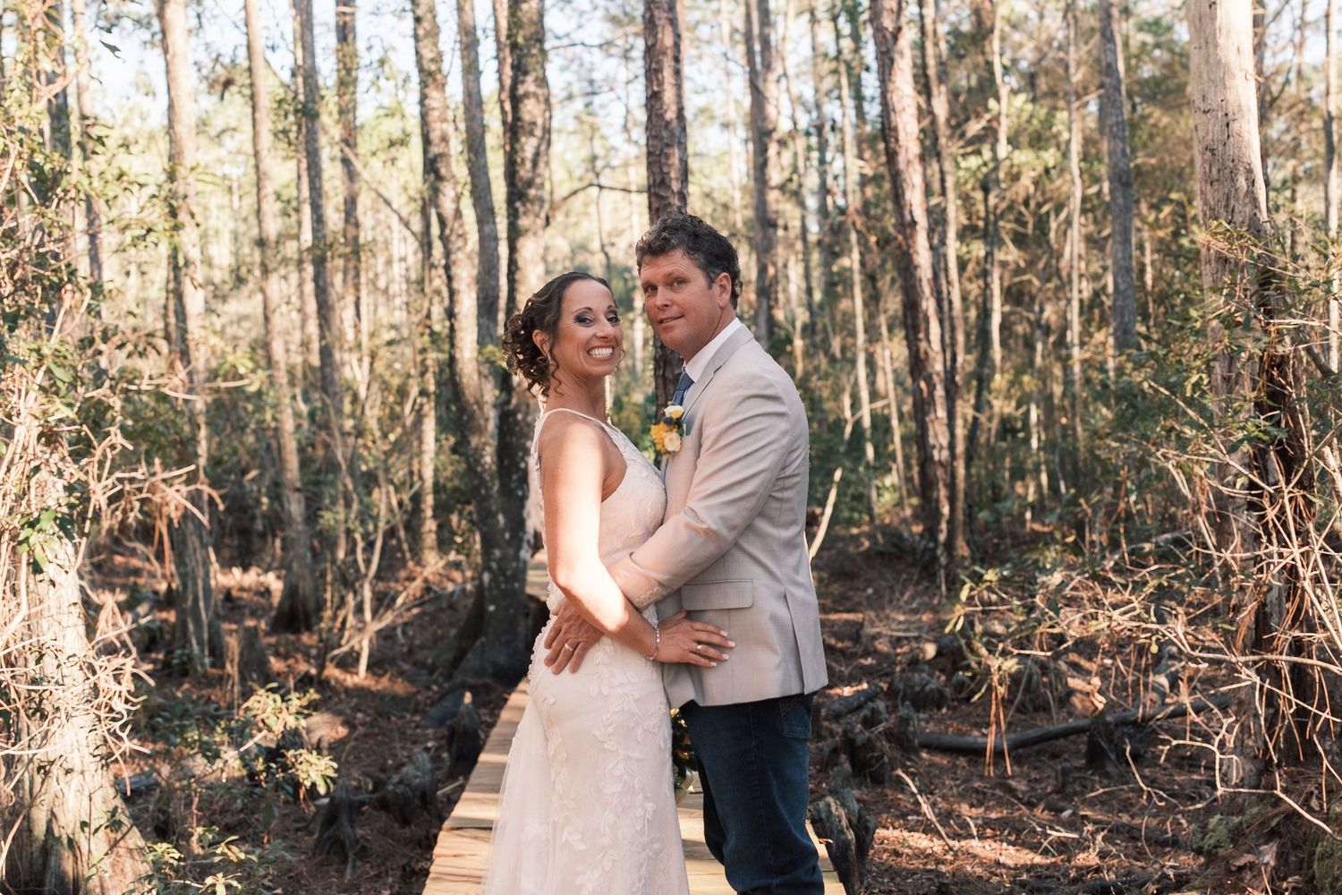 Bride and groom embrace tenderly on a wooden bridge surrounded by a sunlit forest.