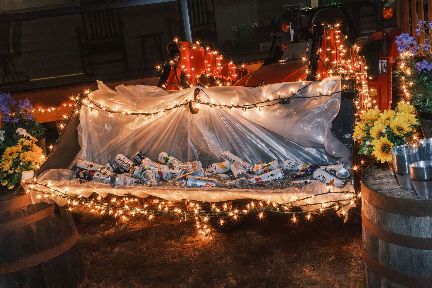 Beer cans displayed under fairy lights and sheer fabric with sunflowers at wedding reception.