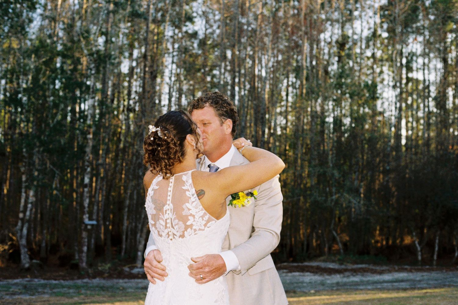 Bride and groom sharing first kiss outdoors with tall pine trees in background at wedding.