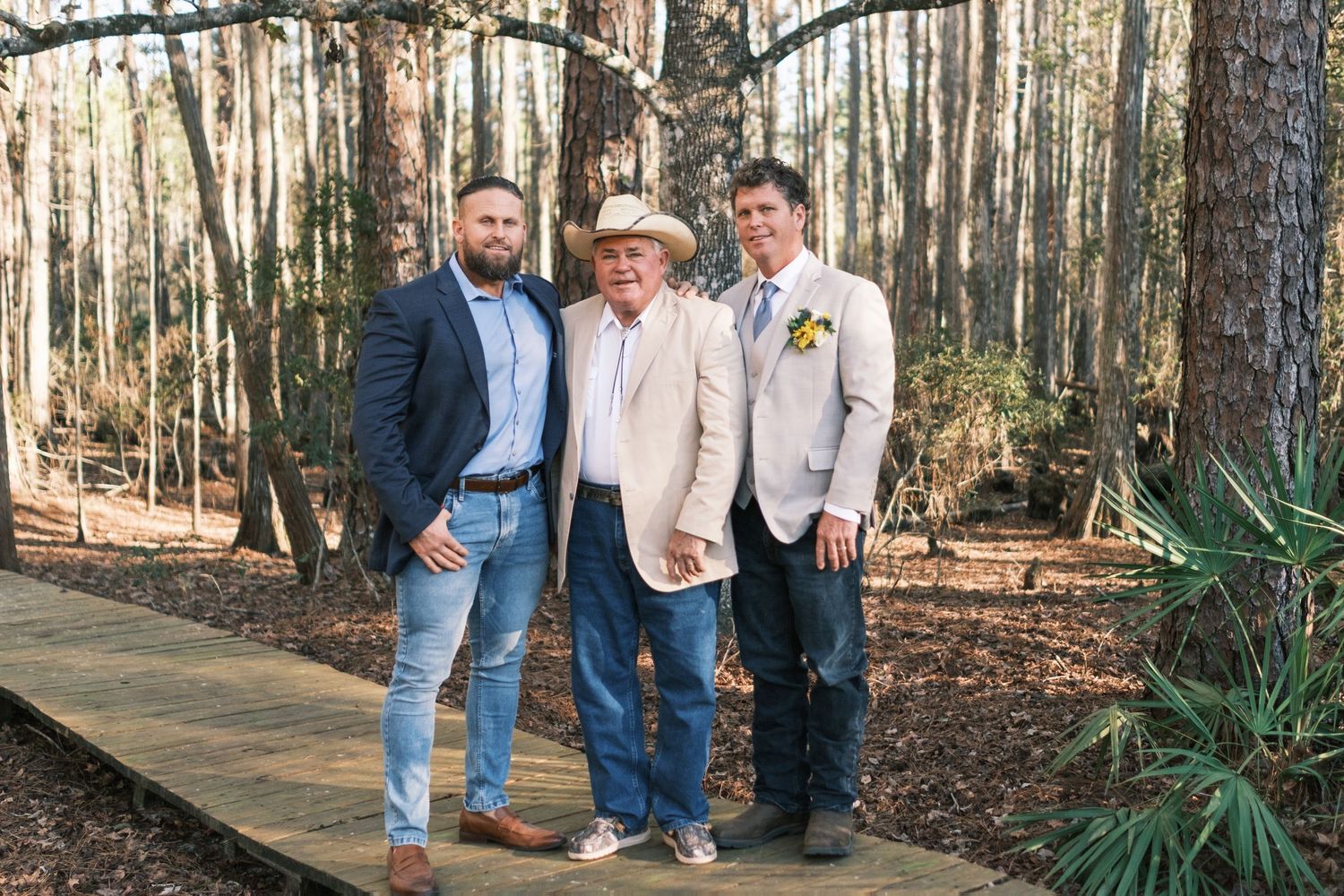 Three men in suits posing together on a wooden boardwalk surrounded by pine trees.