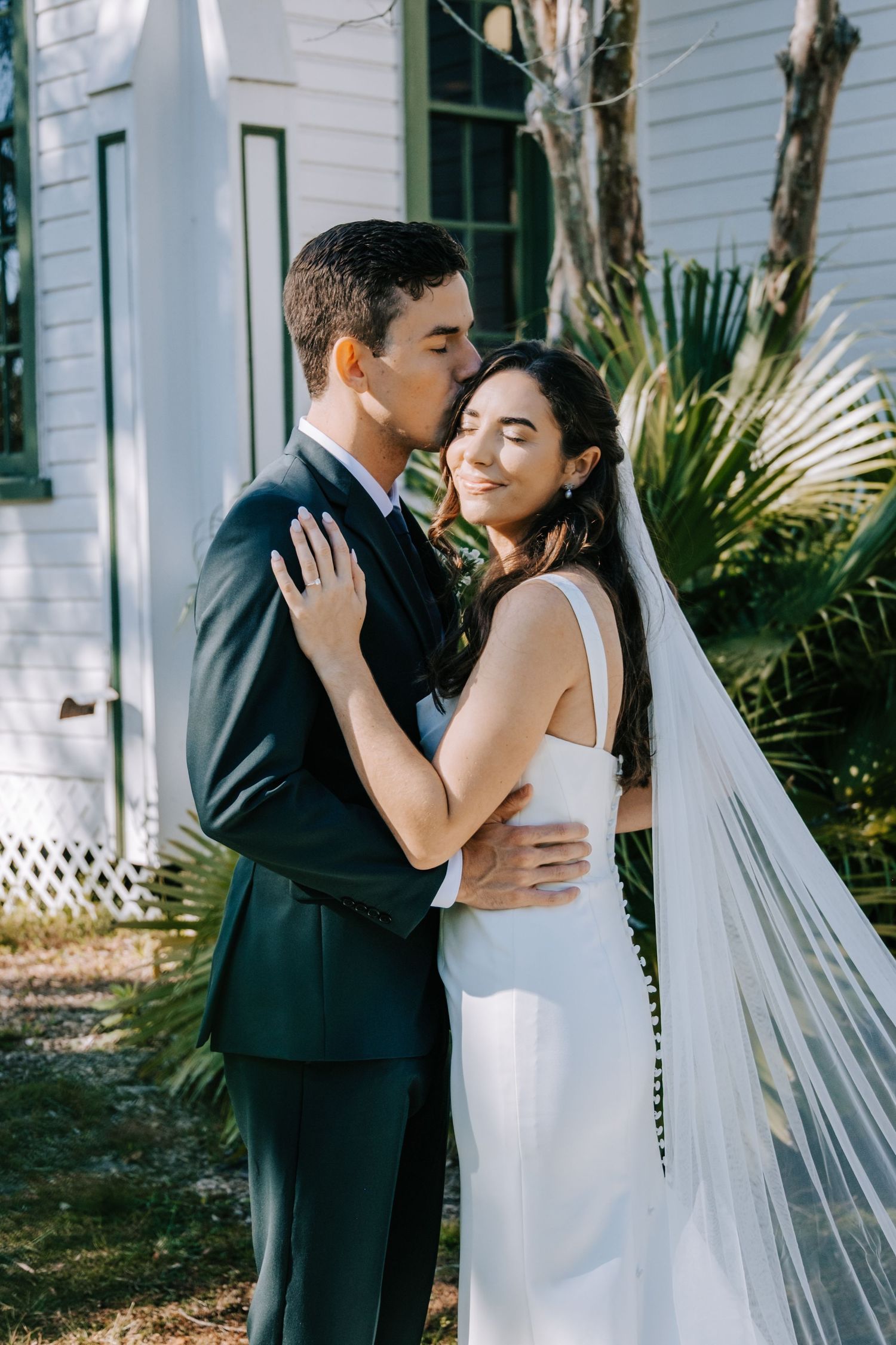 A romantic wedding portrait of a couple embracing outdoors near a white building with palm trees.