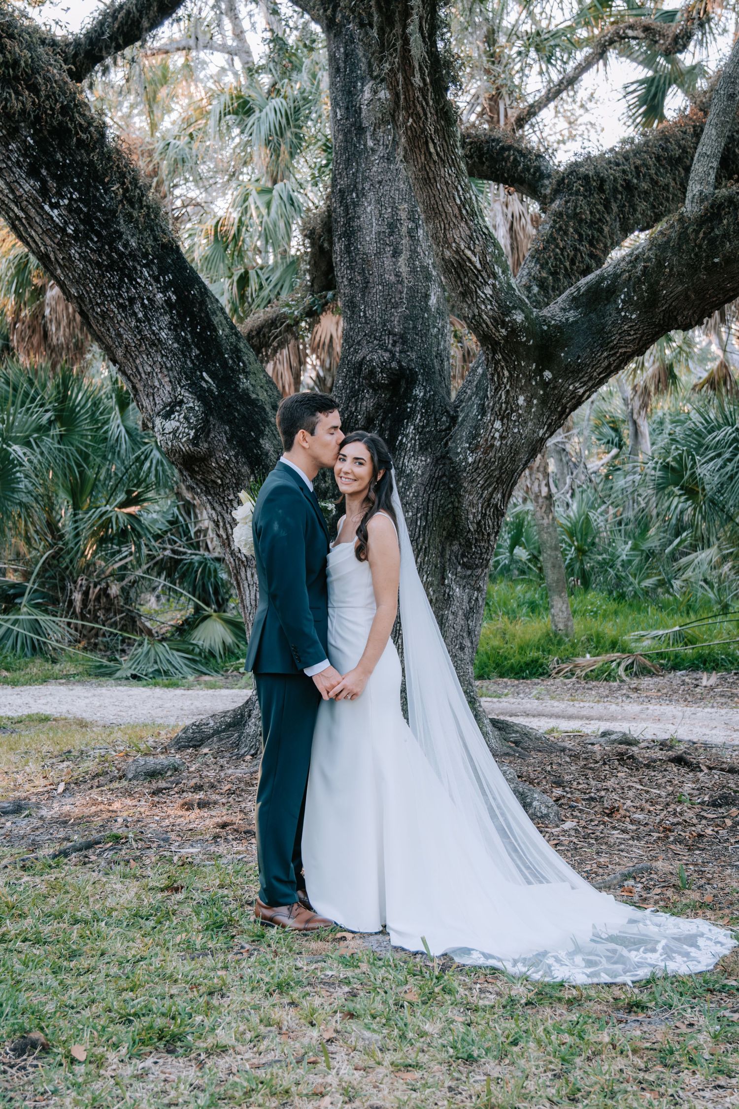 Wedding couple poses romantically under majestic oak trees in an outdoor garden setting.