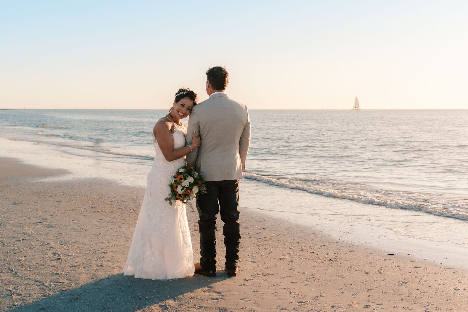Bride and groom stand arm in arm facing ocean at sunset, sailboat visible on horizon.