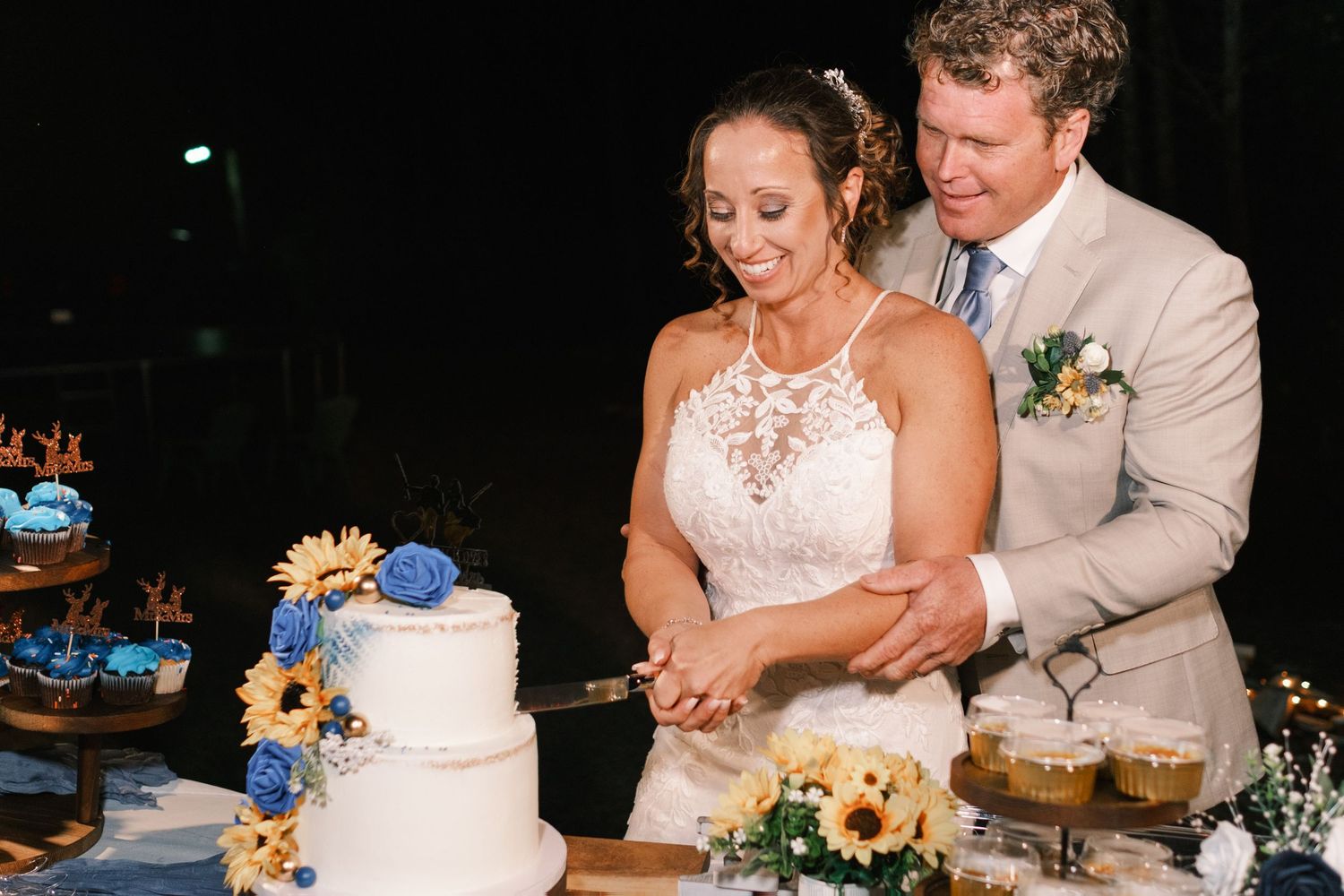 Bride and groom cut white wedding cake decorated with blue roses and sunflowers at night reception.