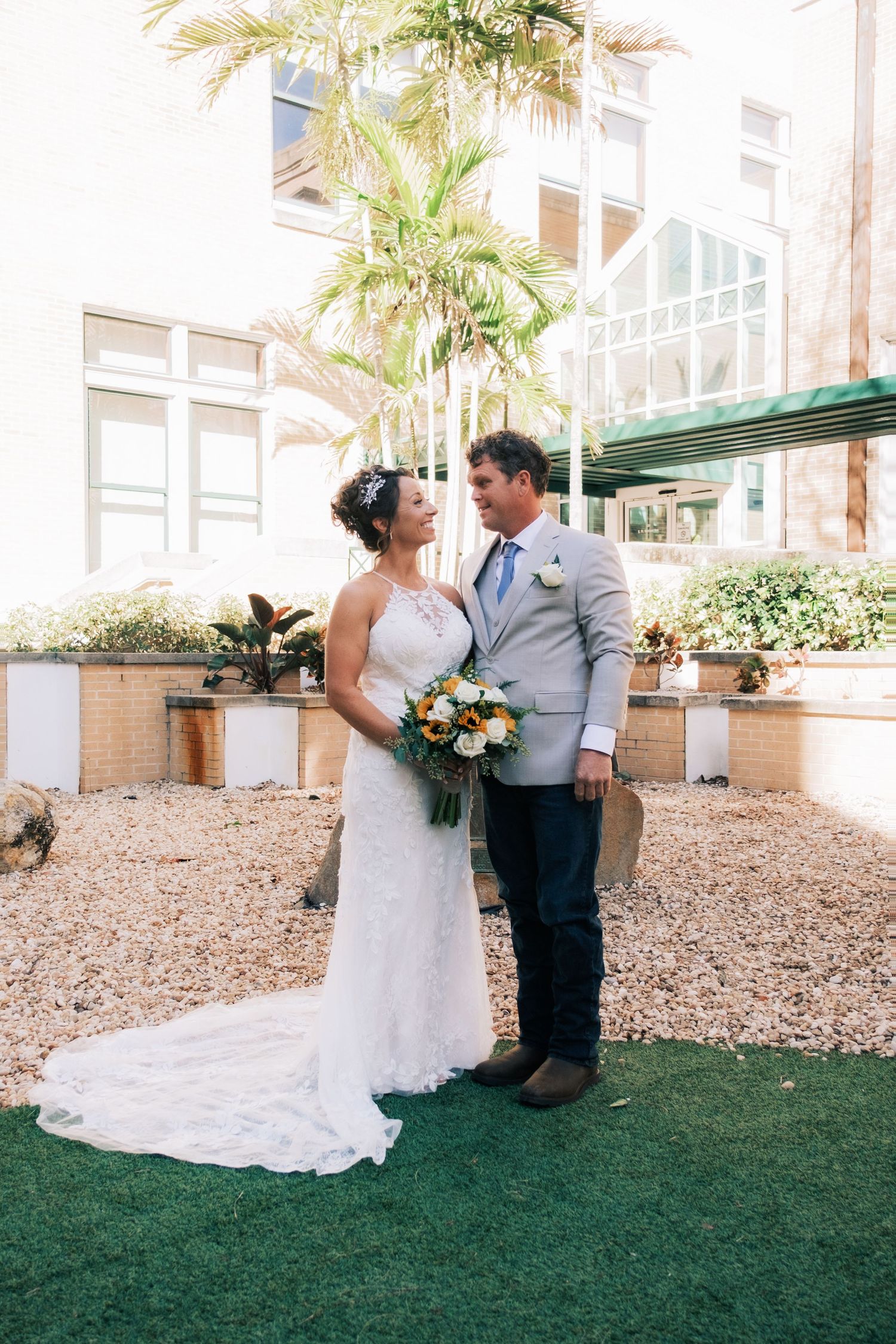 Bride and groom stand together in a sunlit courtyard, she holds a bouquet, both smiling at each other.