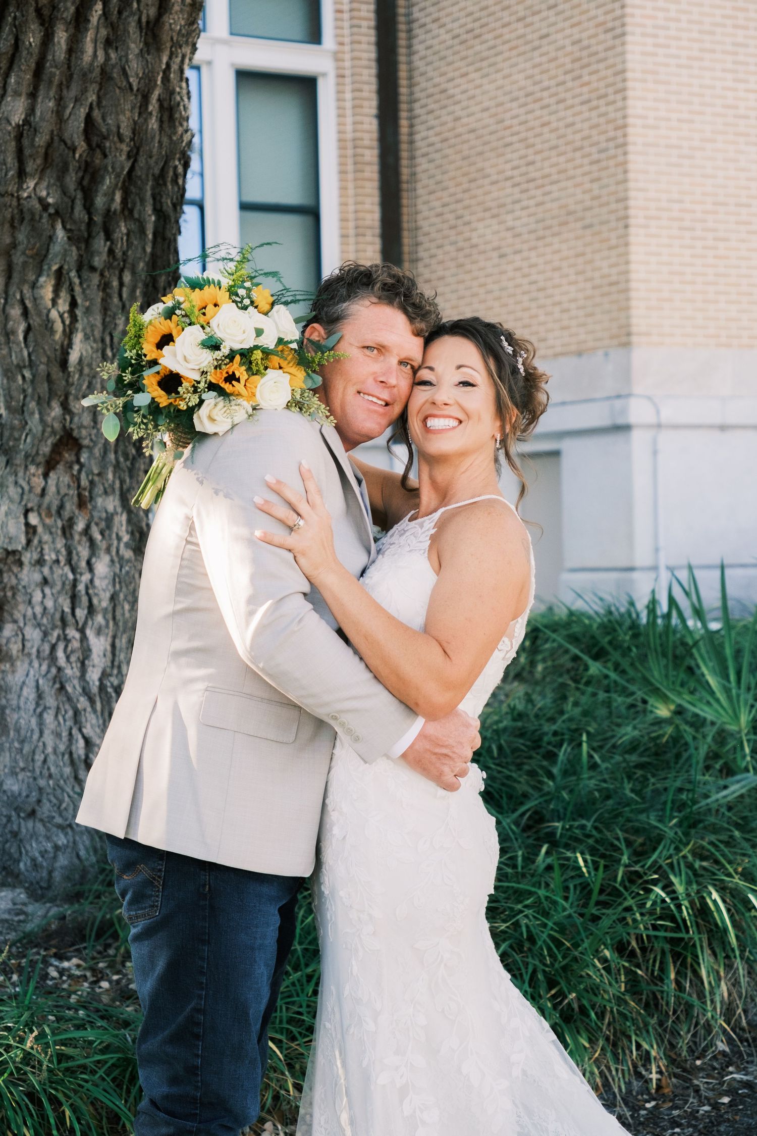 Smiling bride and groom embrace near a large tree holding a sunflower and white rose bouquet.