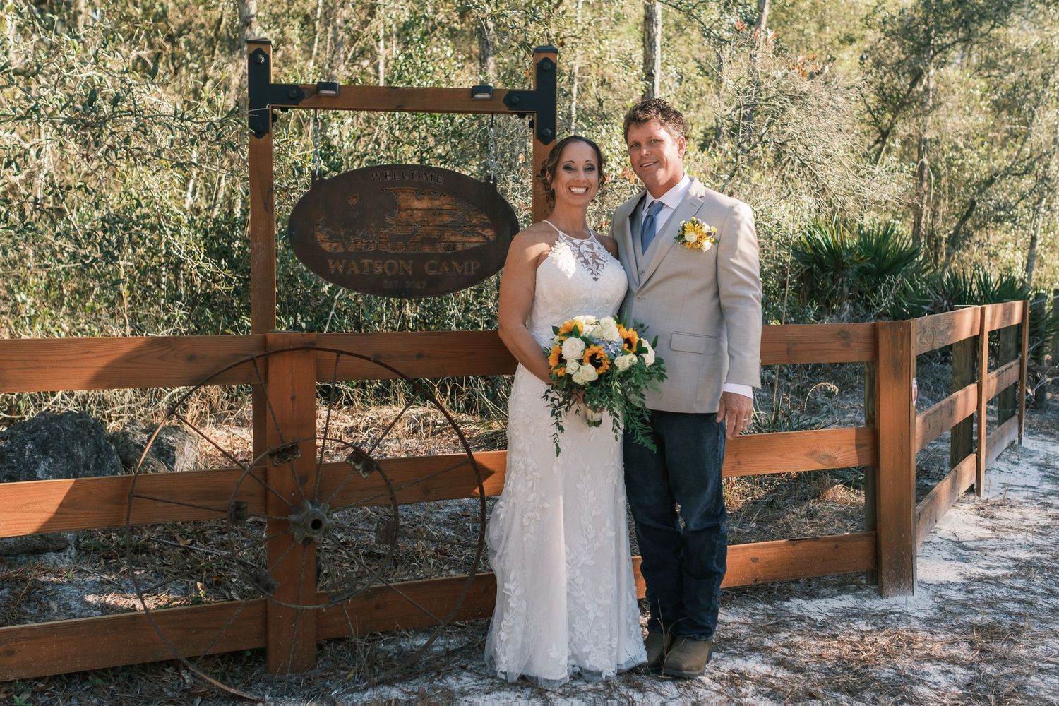 Bride and groom posing together in front of a rustic wooden fence sign surrounded by lush greenery on their wedding day.