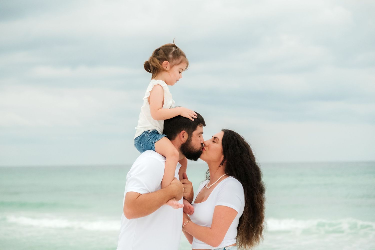 A family shares a tender moment together on a bright sunny beach with turquoise waters in the background.