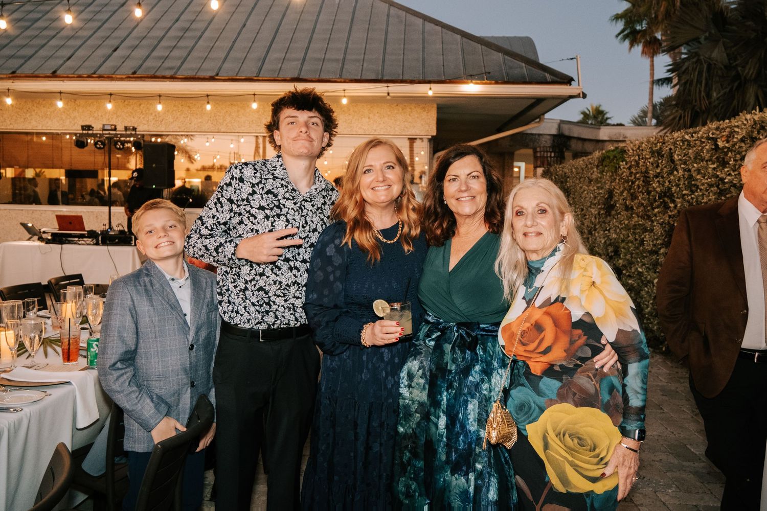 Wedding guests gather outdoors under string lights at a reception during sunset.