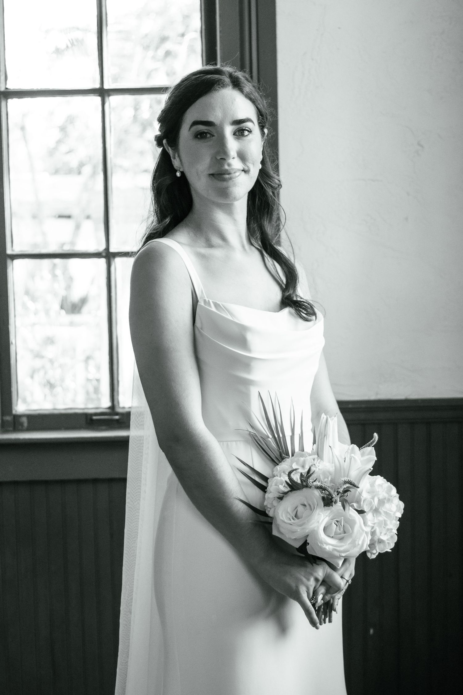 A bride in an elegant white dress holds a bouquet while posing by a window in black and white.