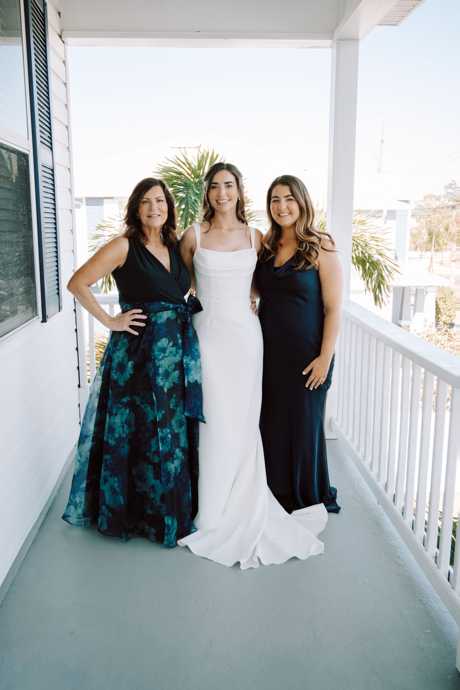Bridesmaids in teal floral dresses pose with bride in white gown on a covered porch.