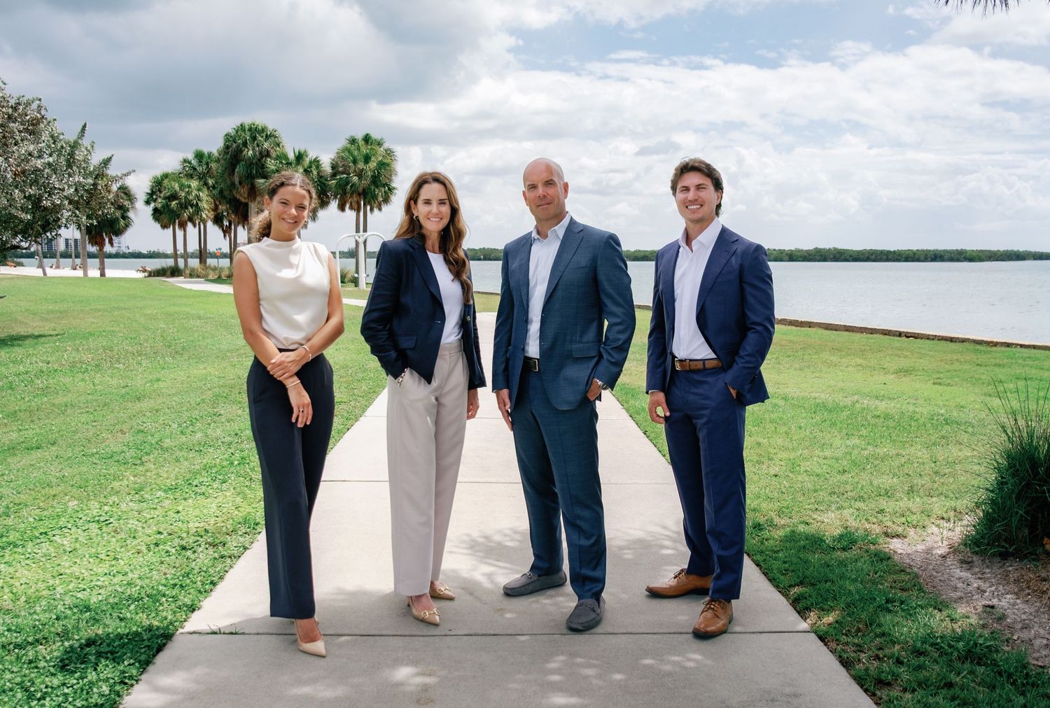 Four real estate professionals posing outdoors near a waterfront park with palm trees and green lawn.