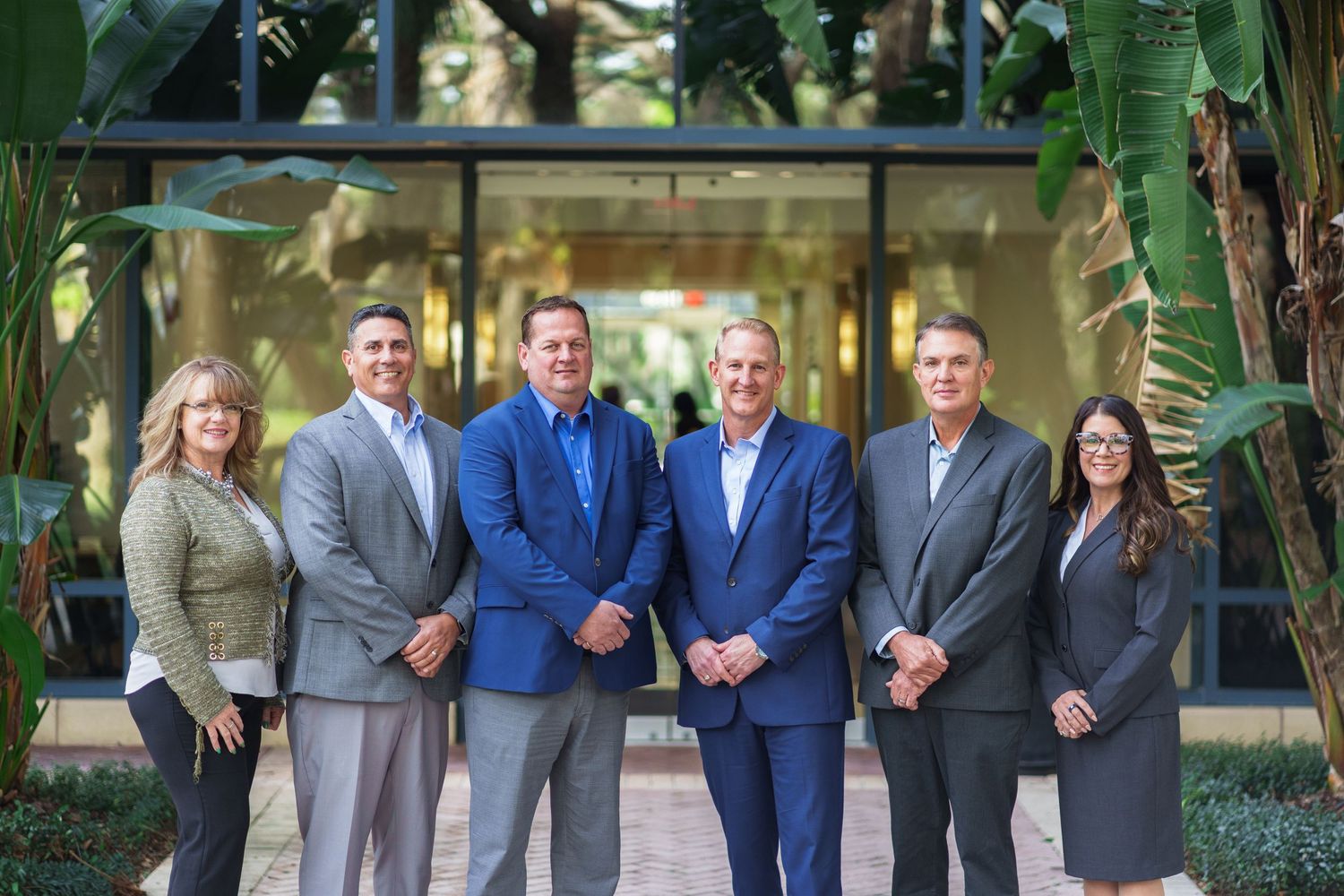 Six business professionals smiling and posing together outdoors in front of a modern glass building surrounded by tropical plants.