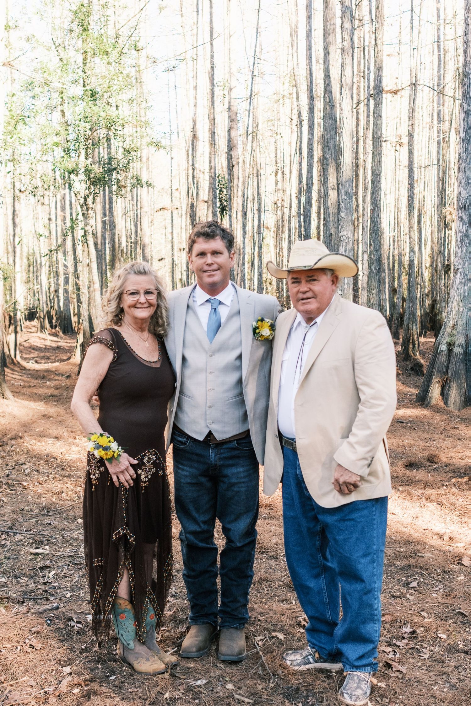 Groom in tan suit and jeans posing with parents in wooded forest setting at wedding.