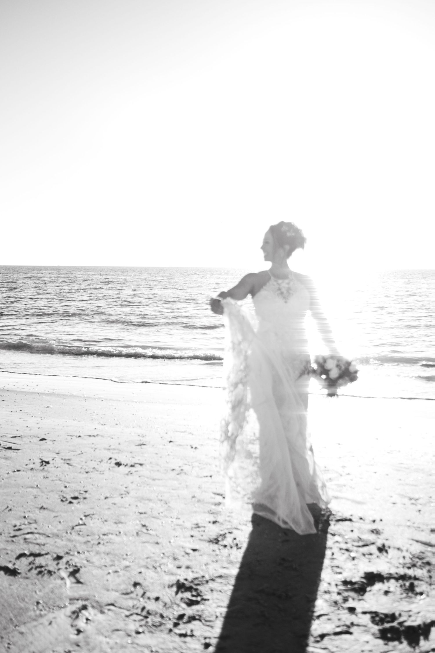 Black and white photo of bride in wedding gown holding bouquet on beach at sunset.