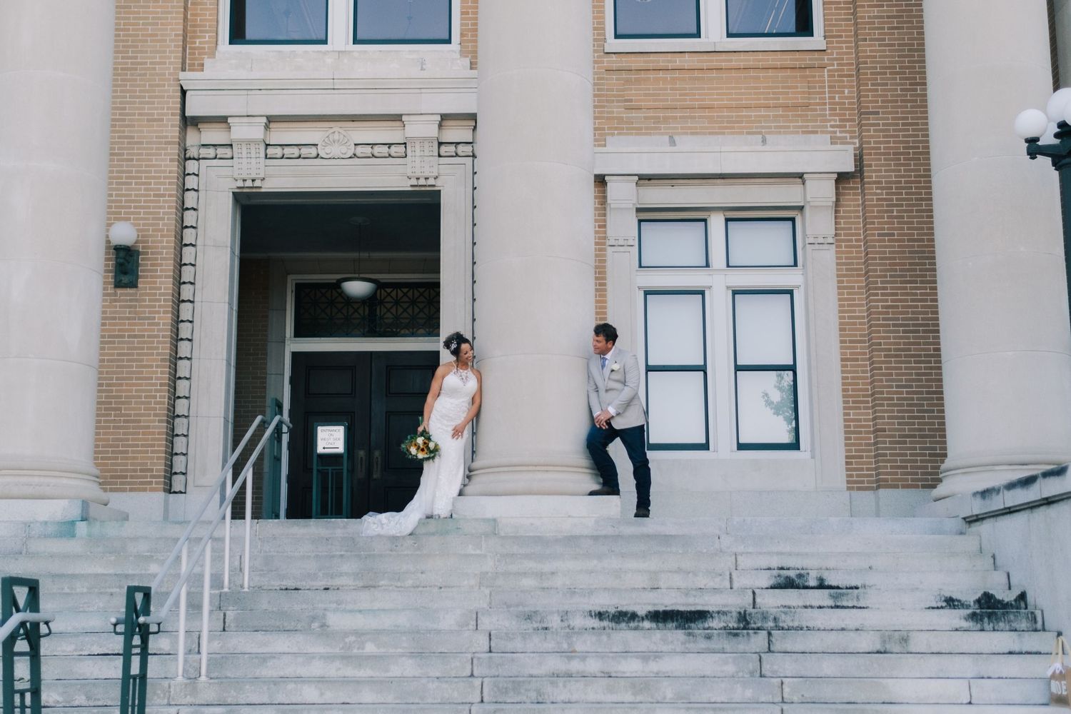 Bride in white gown and groom in gray suit pose on steps of elegant brick building on wedding day.