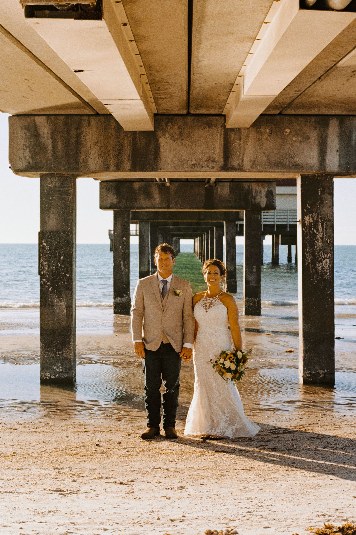 Bride and groom posing together under a concrete pier on the beach.