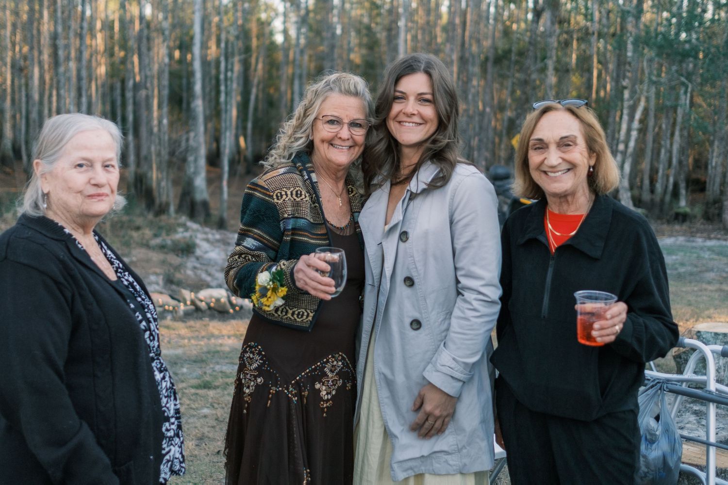 Four women smile together at an outdoor evening wedding reception surrounded by tall trees.