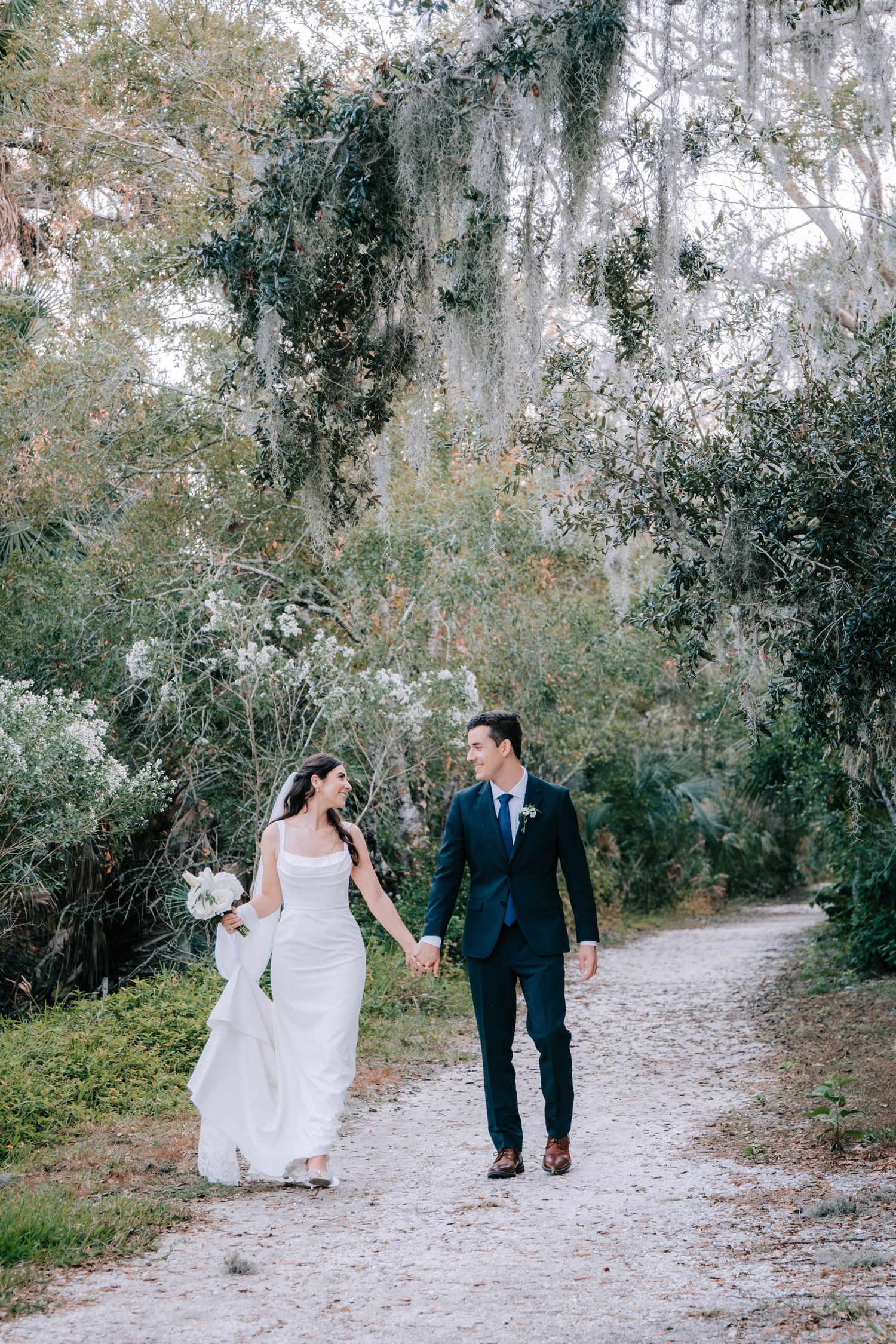 Newlyweds stroll hand in hand down a tree-lined path with Spanish moss overhead.