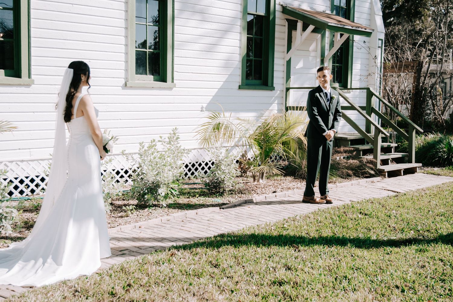 A bride in a white dress walks gracefully across a lawn in front of a white house with green trim.