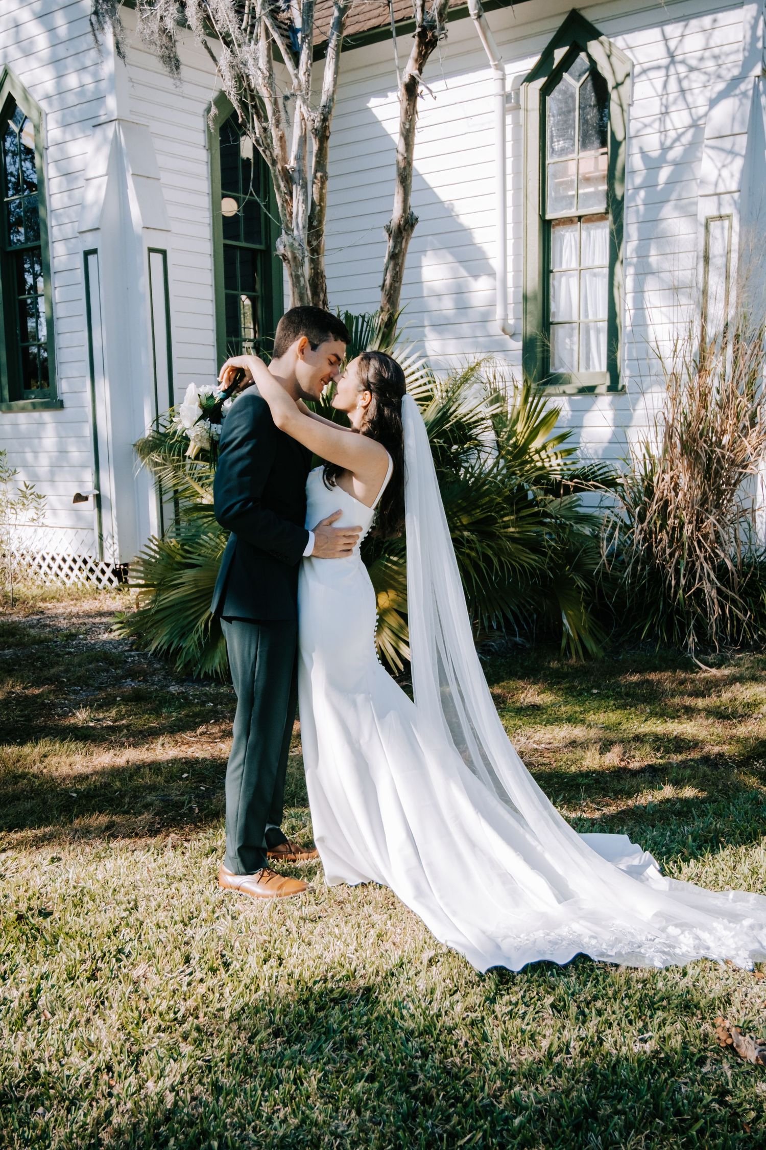 A romantic embrace between newlyweds outside a white chapel with flowing veil and greenery.
