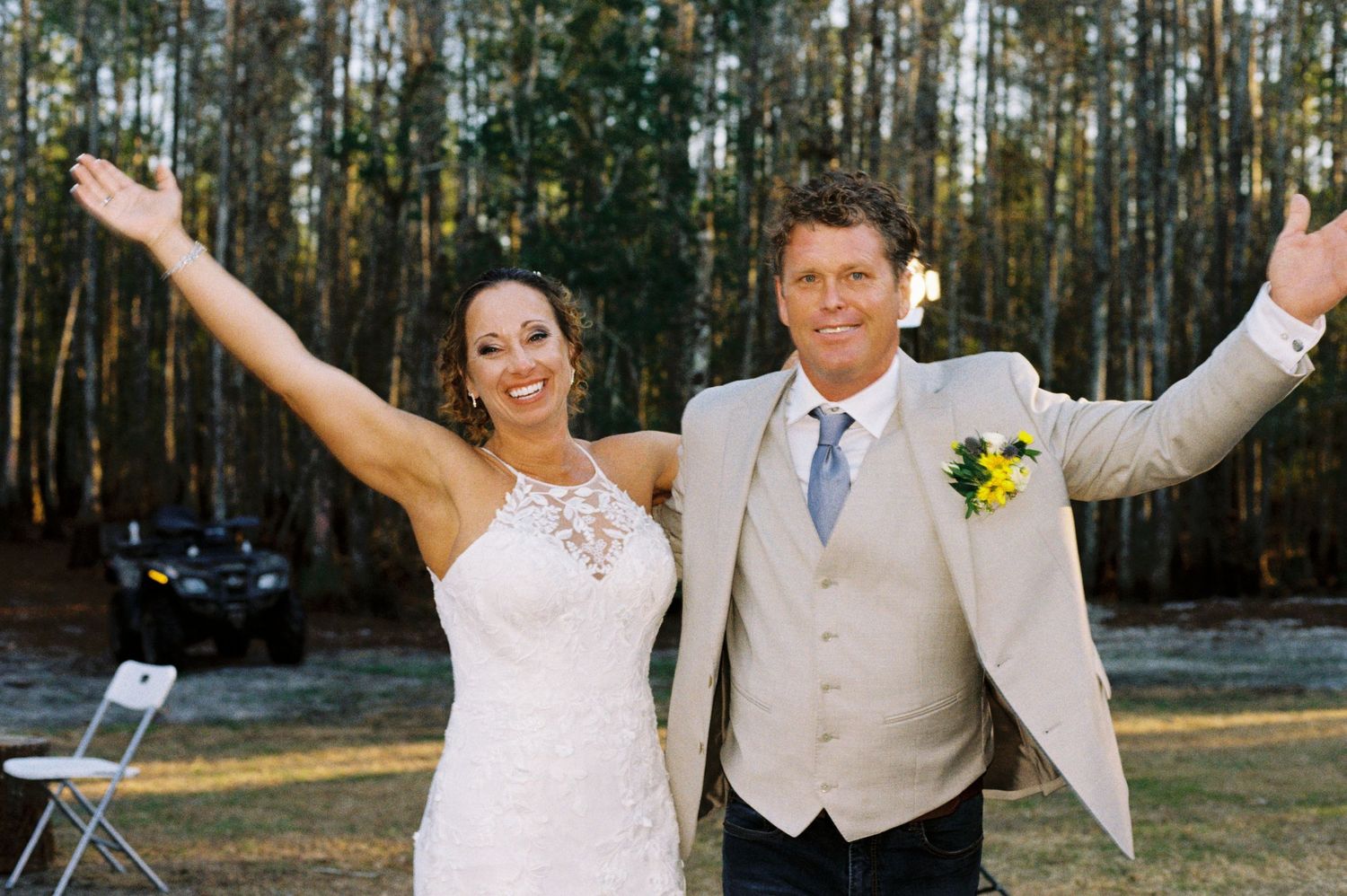 Joyful bride and groom celebrating with arms raised wide at outdoor forest wedding.