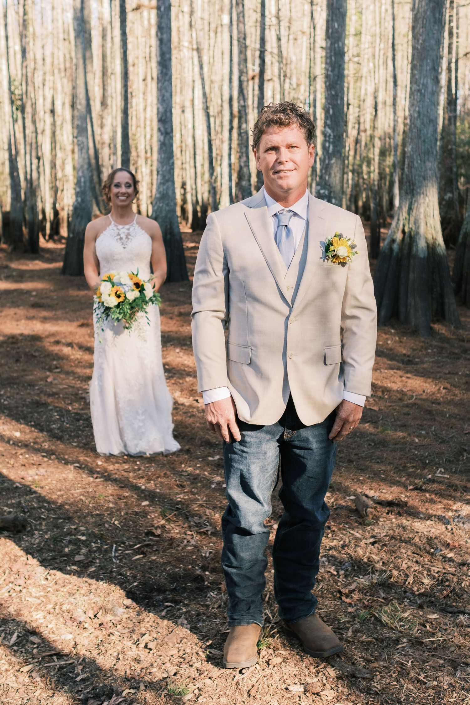 Groom in beige blazer and jeans stands in pine forest as bride in white gown holds sunflower bouquet behind him.
