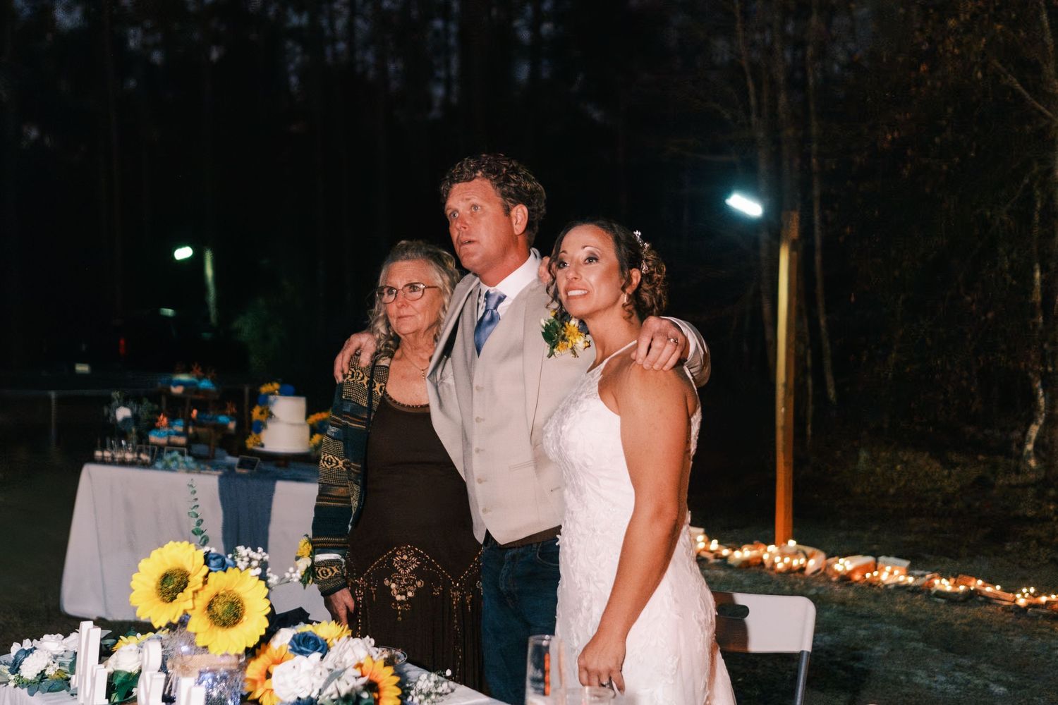 Bride and groom pose with family member at nighttime outdoor wedding reception table.