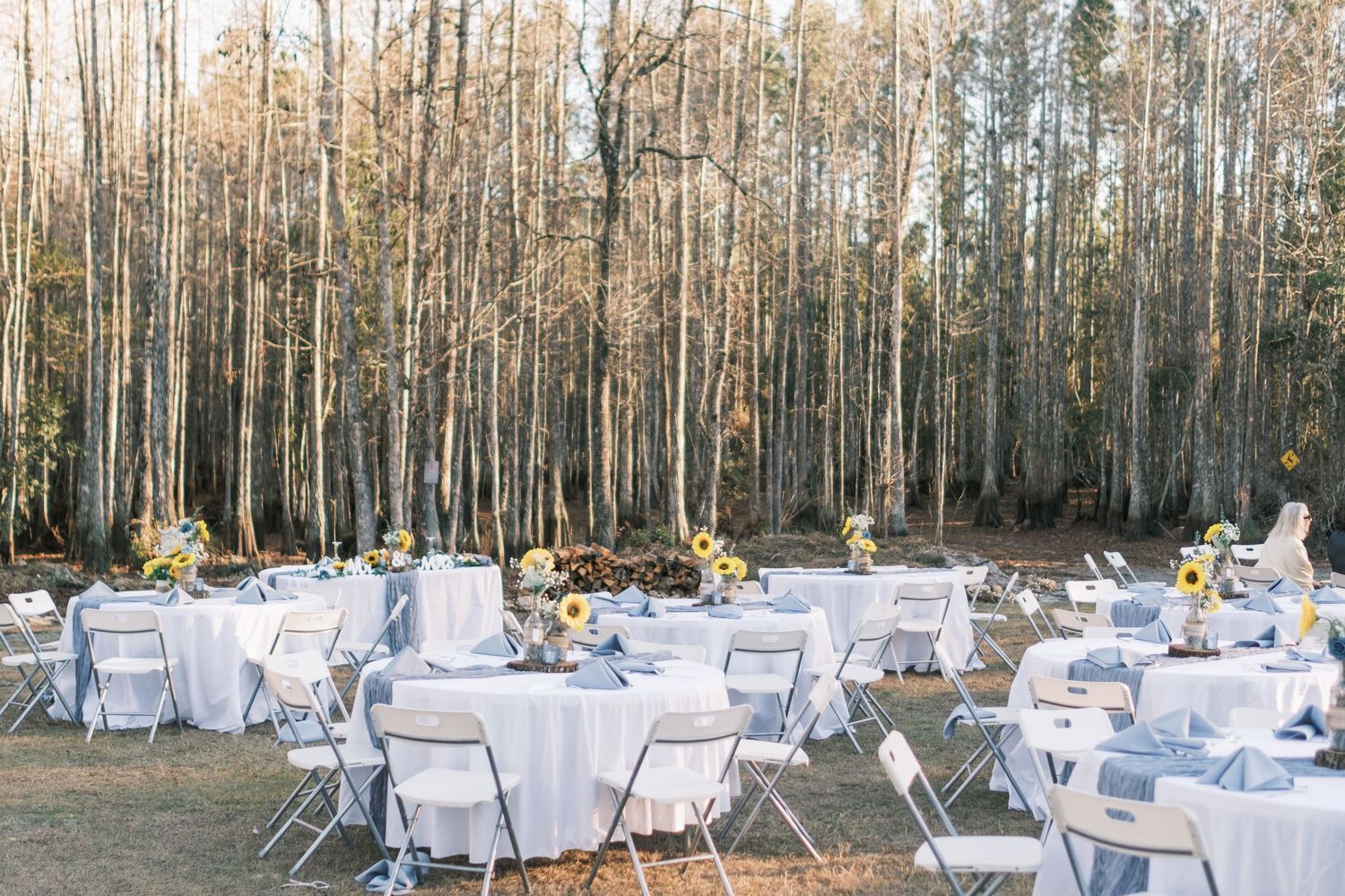 Elegant outdoor wedding reception with round tables, white chairs, and sunflower centerpieces among tall trees.