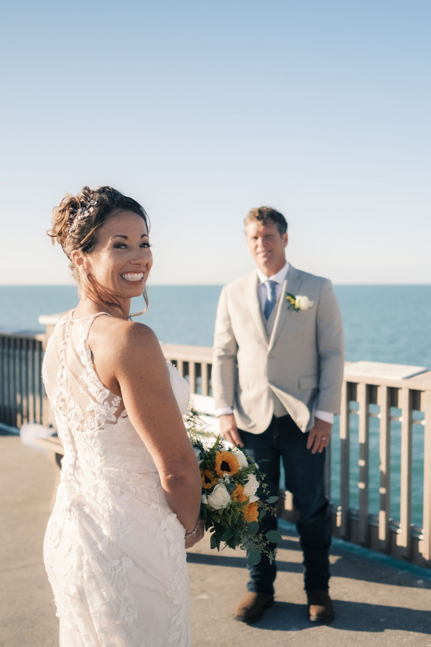 Smiling bride in lace gown holds bouquet on pier with groom in tan suit and ocean backdrop.