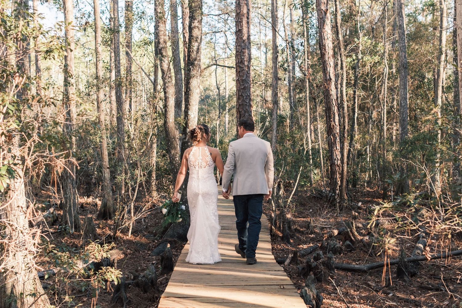 Bride and groom walking hand in hand on a wooden boardwalk through a sunlit forest.
