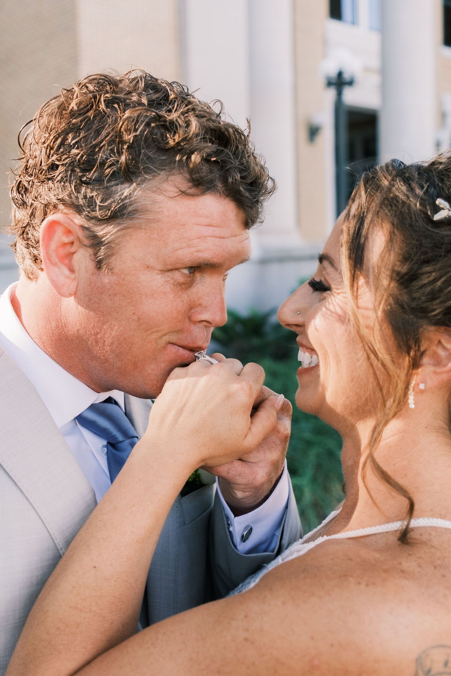 Groom kissing bride's hand while she laughs joyfully during their wedding day portrait session.