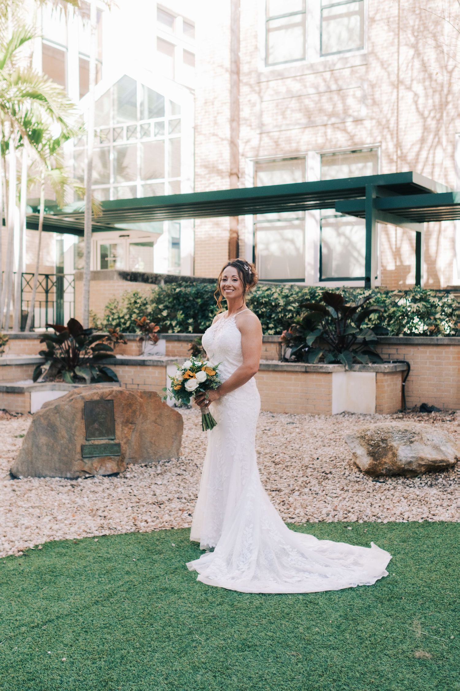 Bride in white lace gown holding bouquet poses in outdoor garden setting.