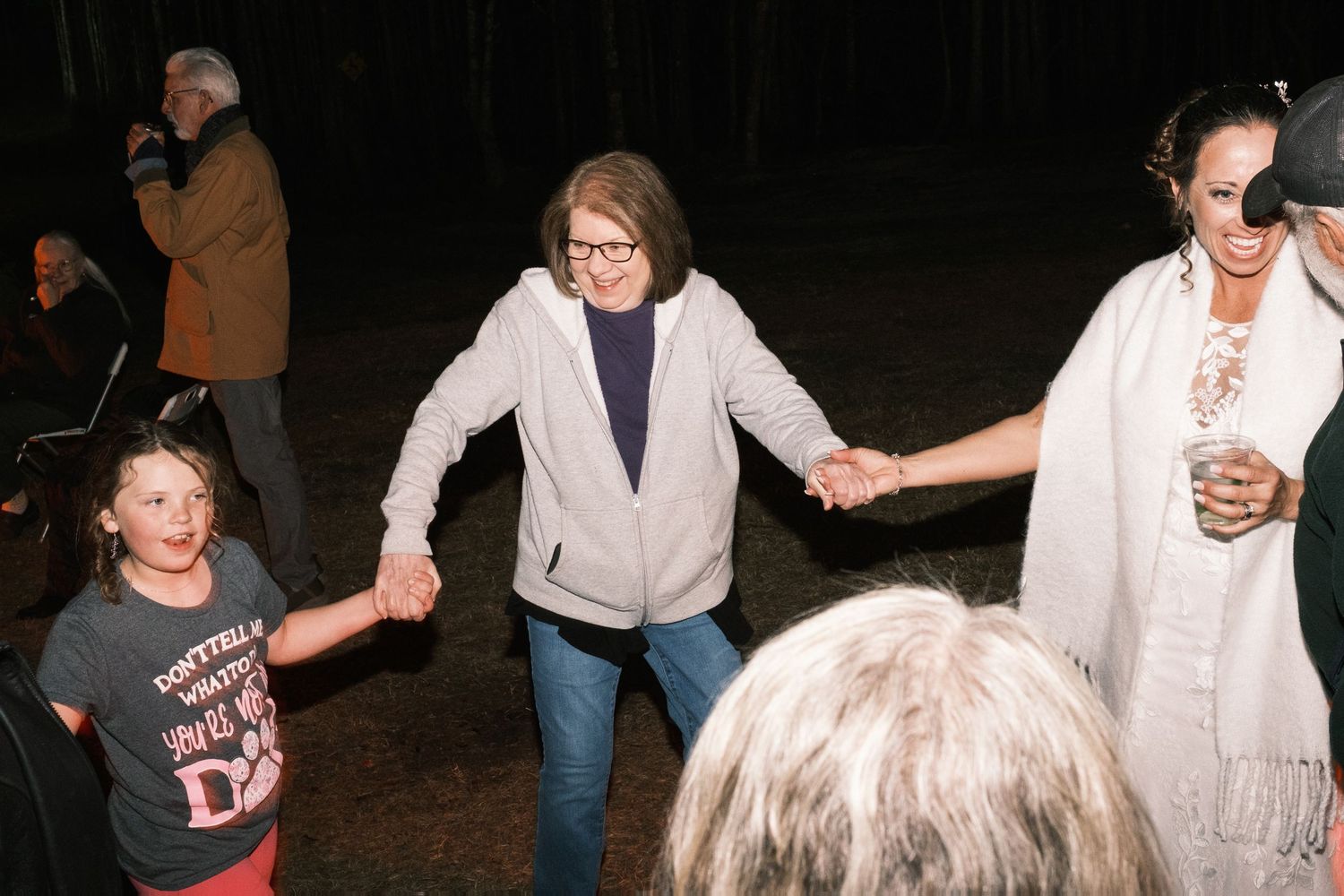 Guests of all ages holding hands and dancing together at an outdoor nighttime wedding reception.