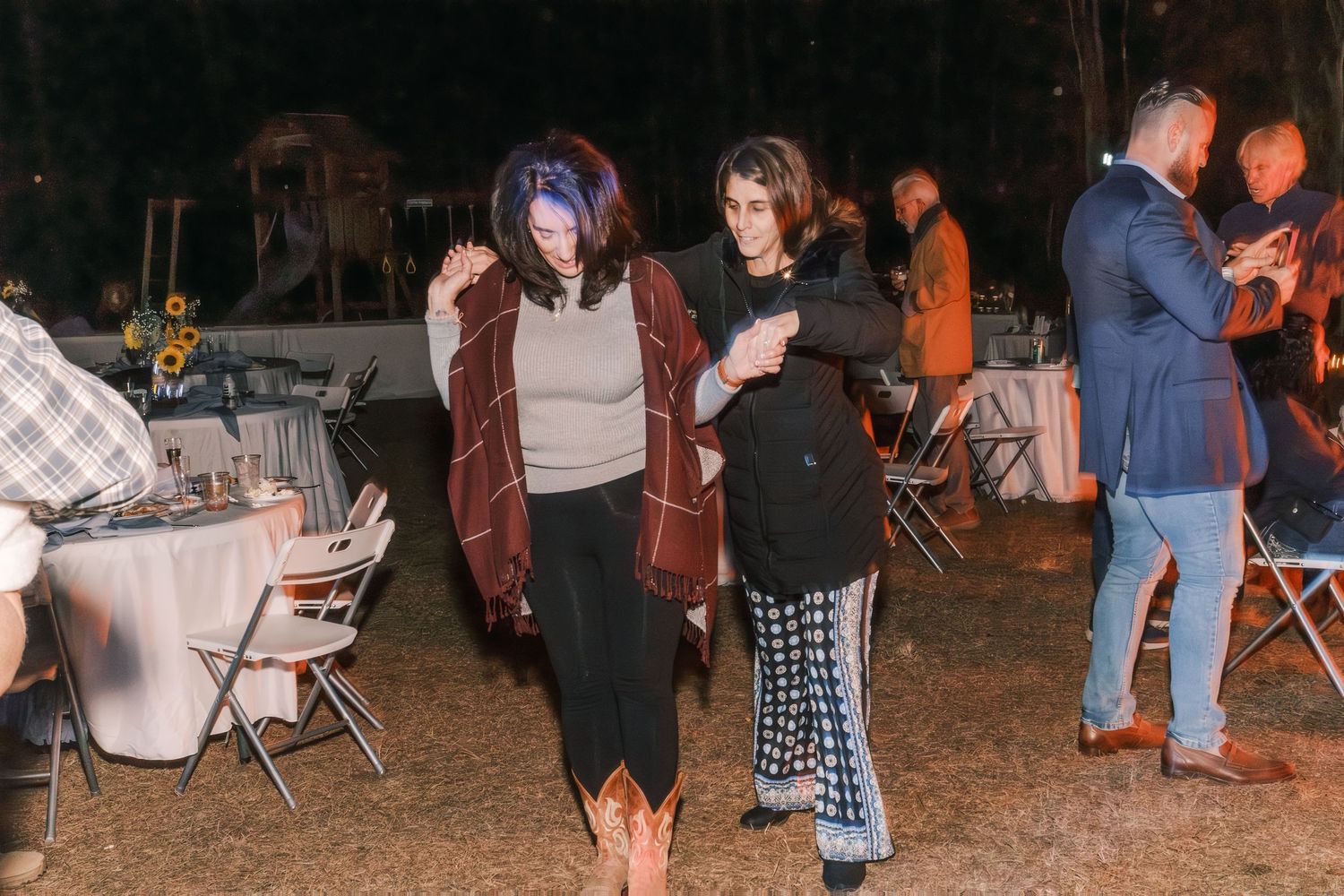 Two women dance together at an outdoor evening wedding reception under string lights.