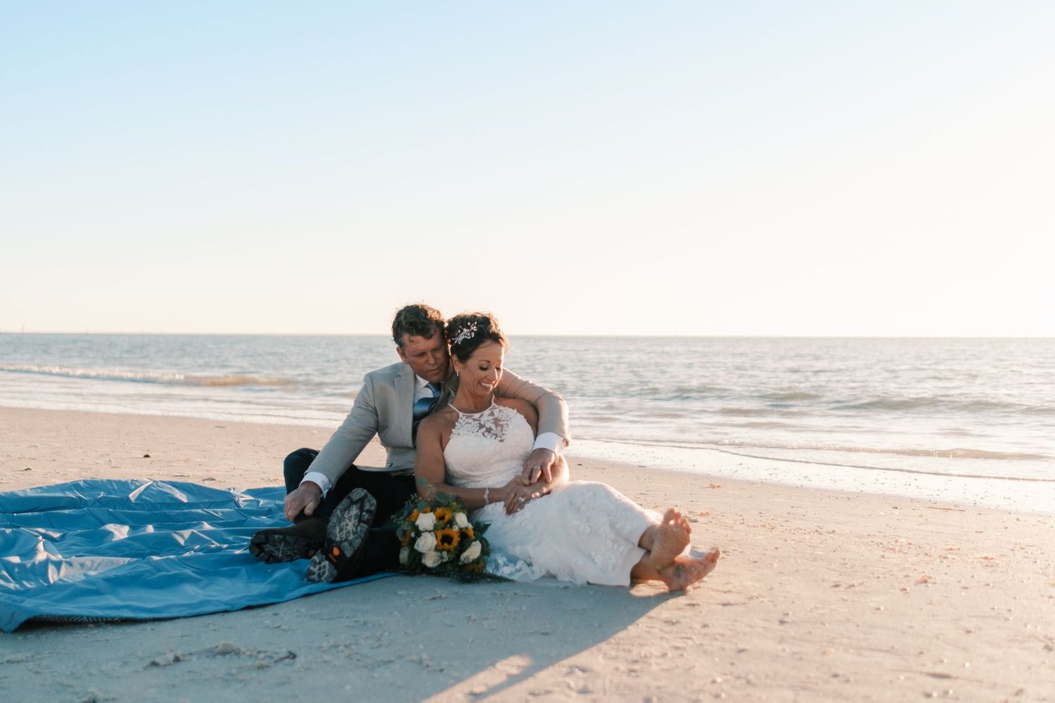 Bride and groom sit together on a blue blanket on the beach, laughing as waves roll in behind them.