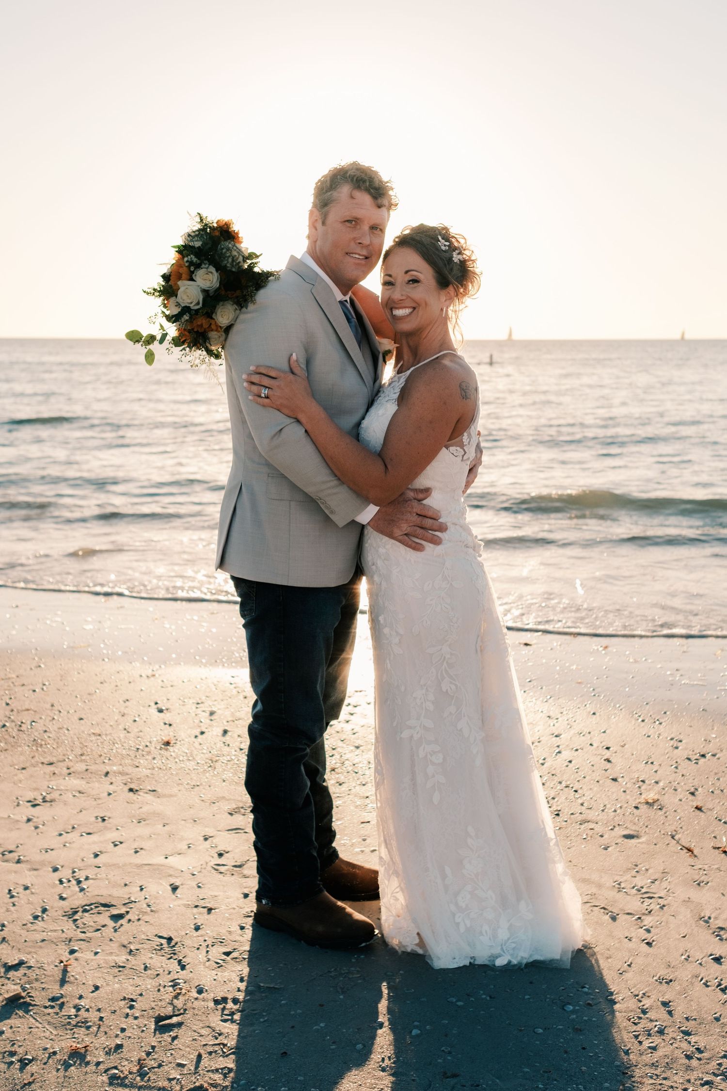 Smiling bride and groom embracing on sandy beach at golden sunset, groom holding floral bouquet.
