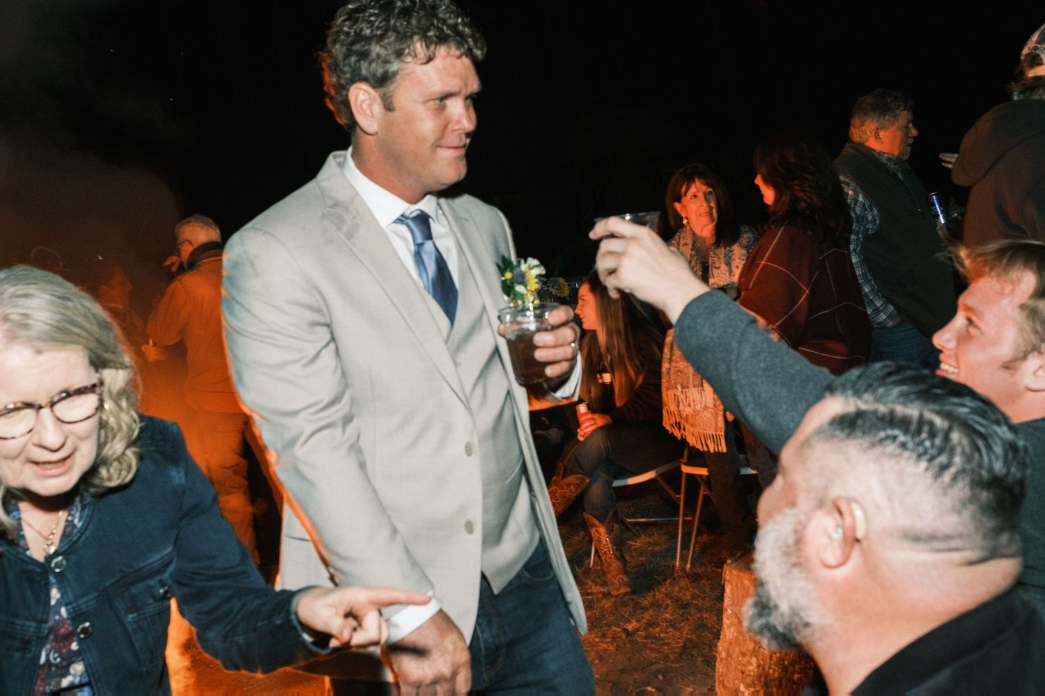 Groom in tan suit mingles with guests holding drinks around an outdoor evening wedding bonfire.