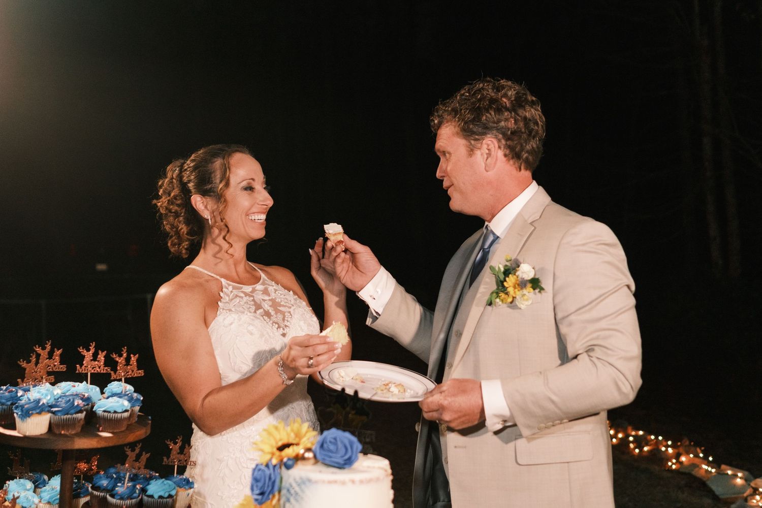 Bride and groom share wedding cake during their outdoor evening reception with blue floral decorations.