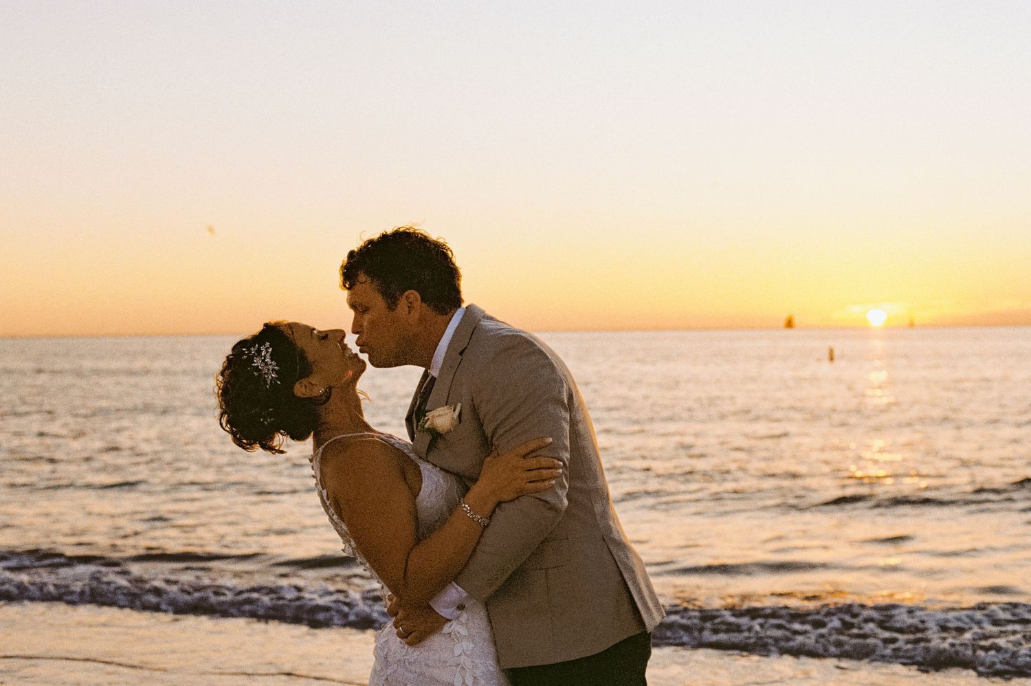 Bride and groom sharing romantic kiss on beach at golden sunset by the ocean.