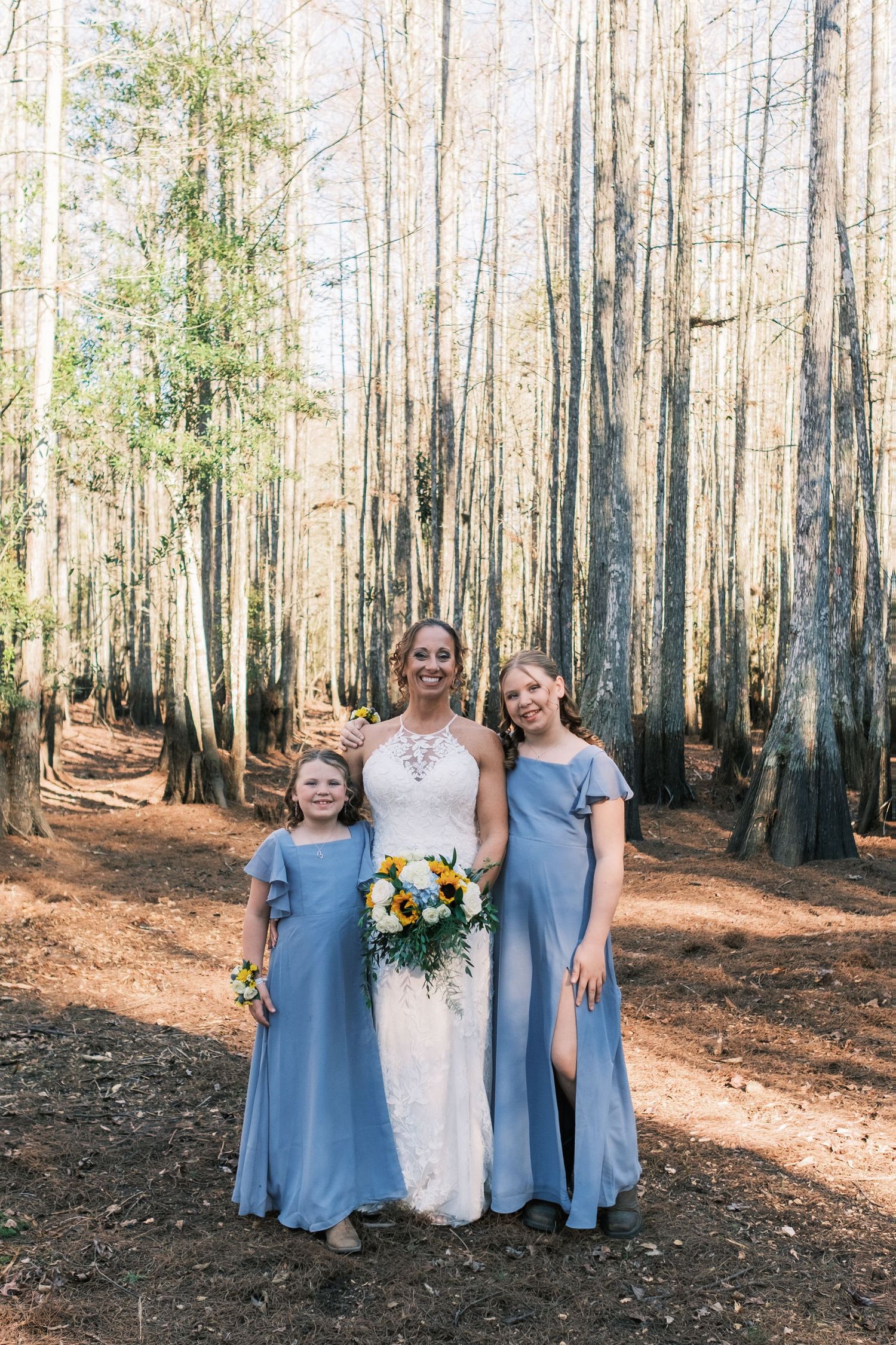 Bride in lace gown poses with bridesmaid and flower girl in blue dresses in forest.