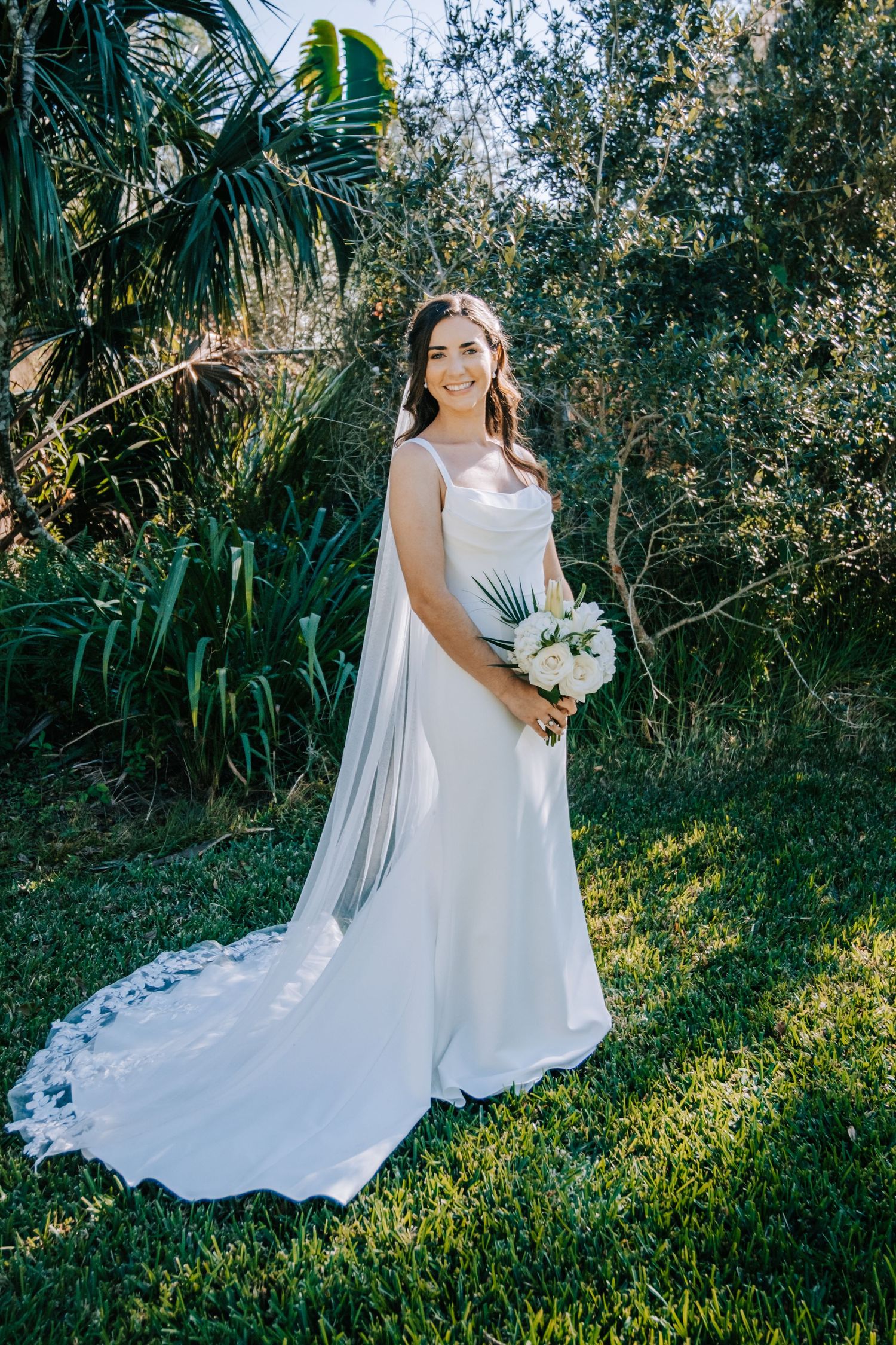 A flowing white wedding dress with a long train photographed in a lush tropical garden setting.