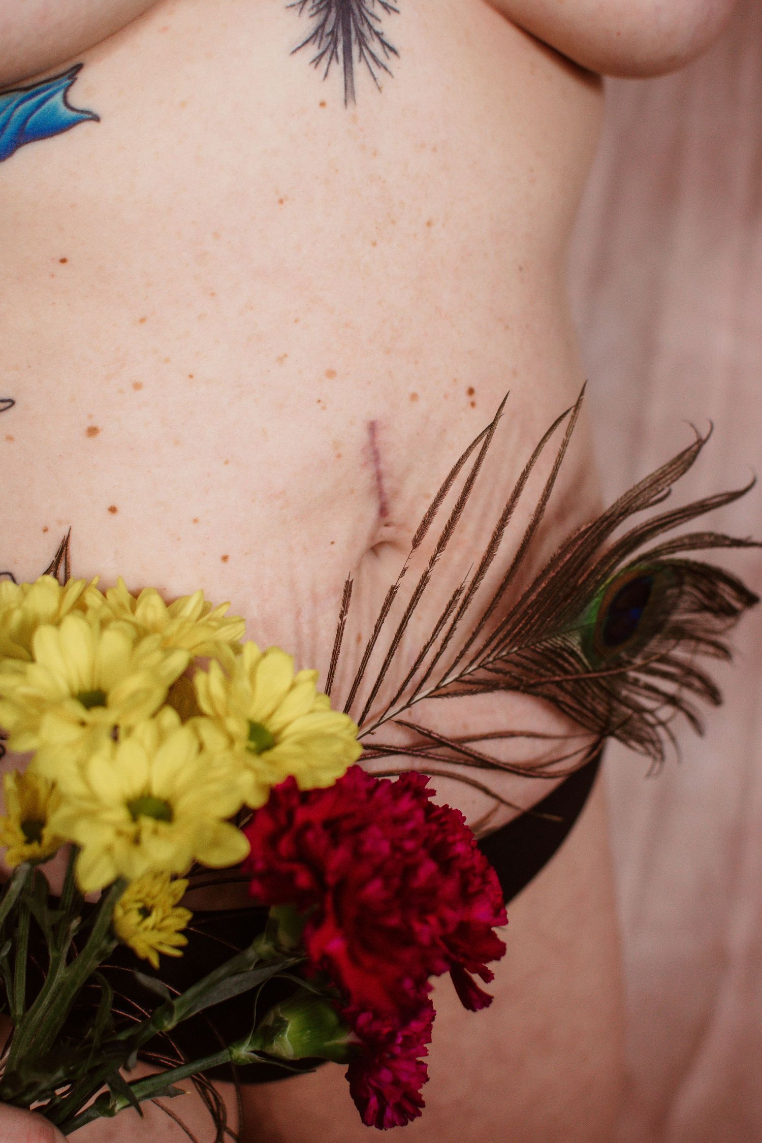 An artistic arrangement of yellow chrysanthemums and deep red carnations with dark feathers against a beige background.