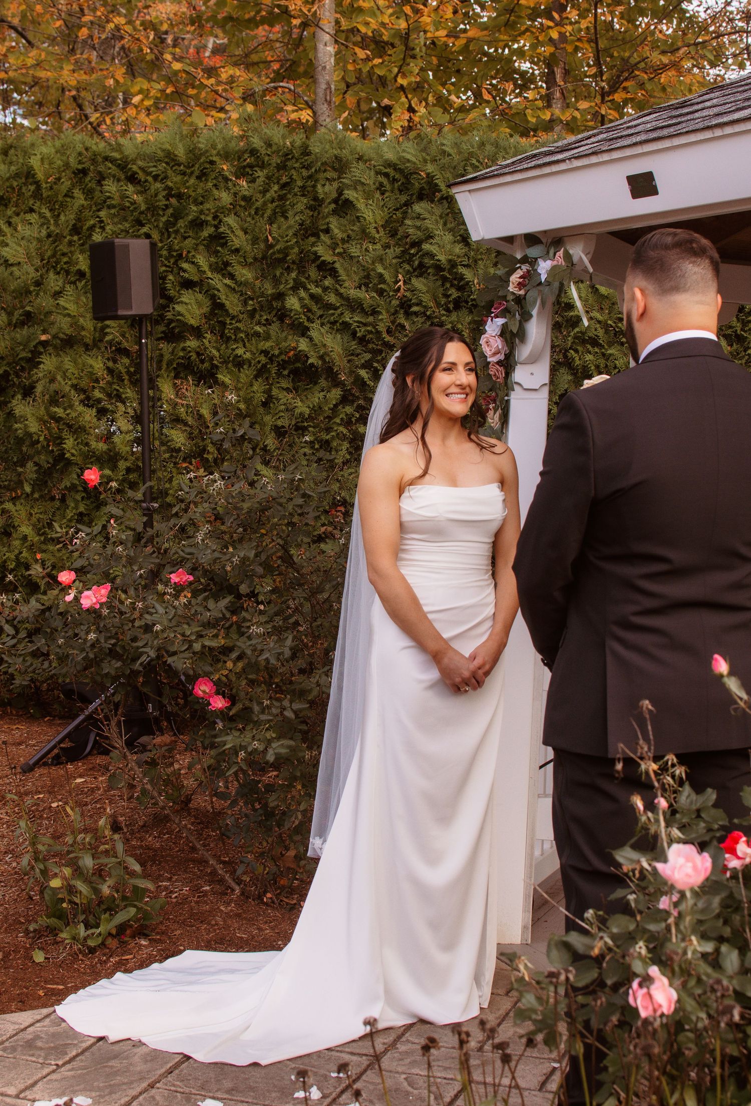 A wedding couple stands together in a garden setting with autumn foliage and pink roses in the background.