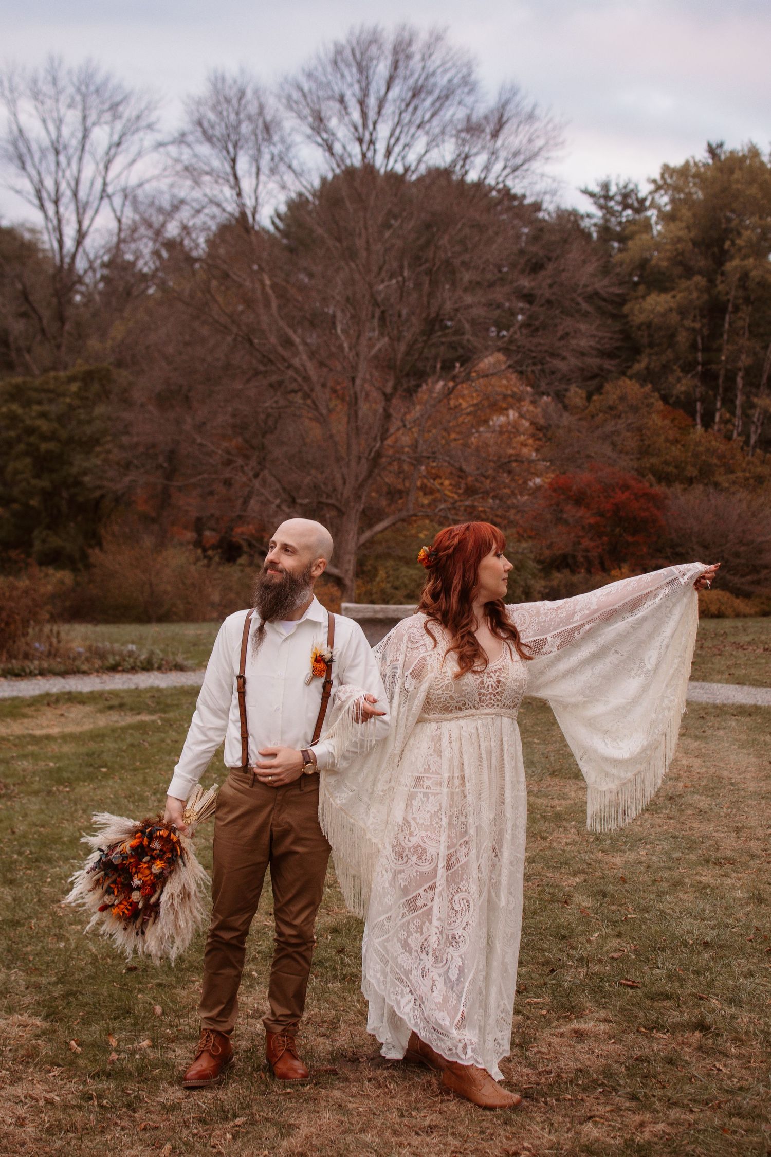 A boho wedding couple poses in a fall landscape with the bride in lace and groom in suspenders holding autumn flowers.