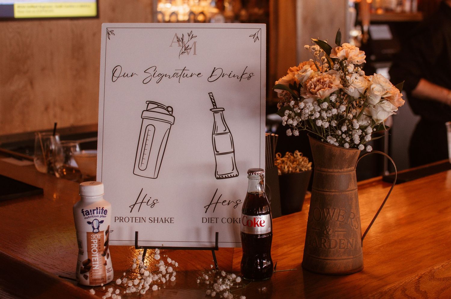 Wedding signature drinks sign displaying his protein shake and her Diet Coke, with floral arrangement on a wooden bar.